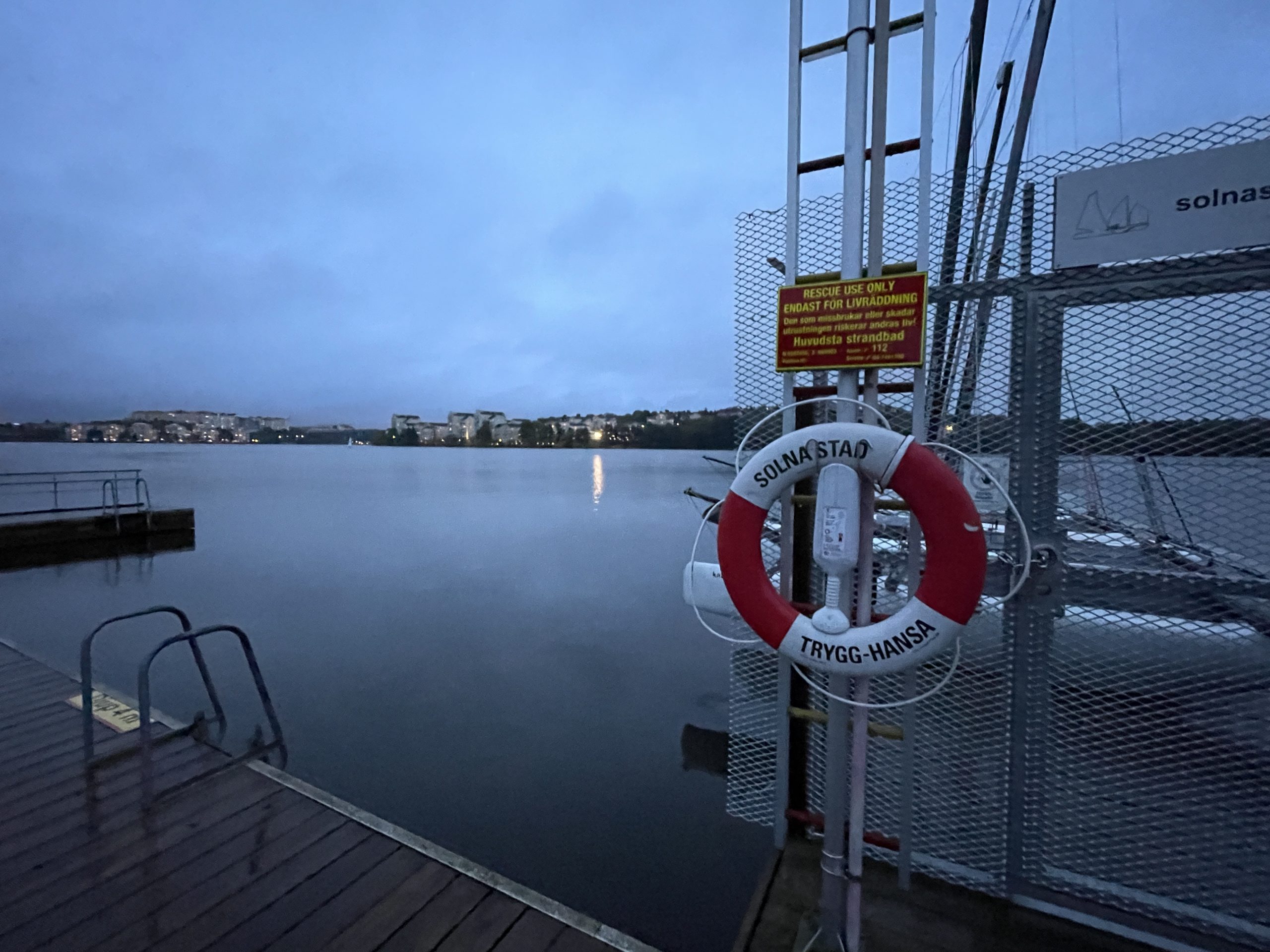 view of lake from the dock with dock in the bottom of the frame and life preserver mounted on metal rack in foreground. Solana, Stockholm Sweden.